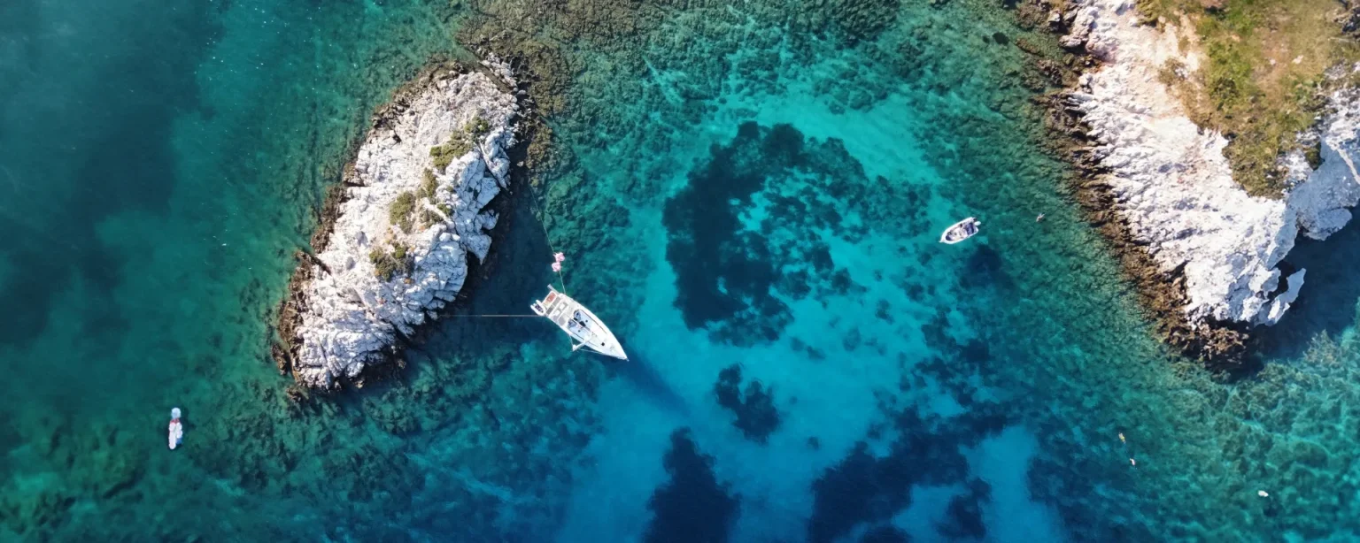 A yacht in turquoise water off a rocky island in the Dodecanese, seen from above