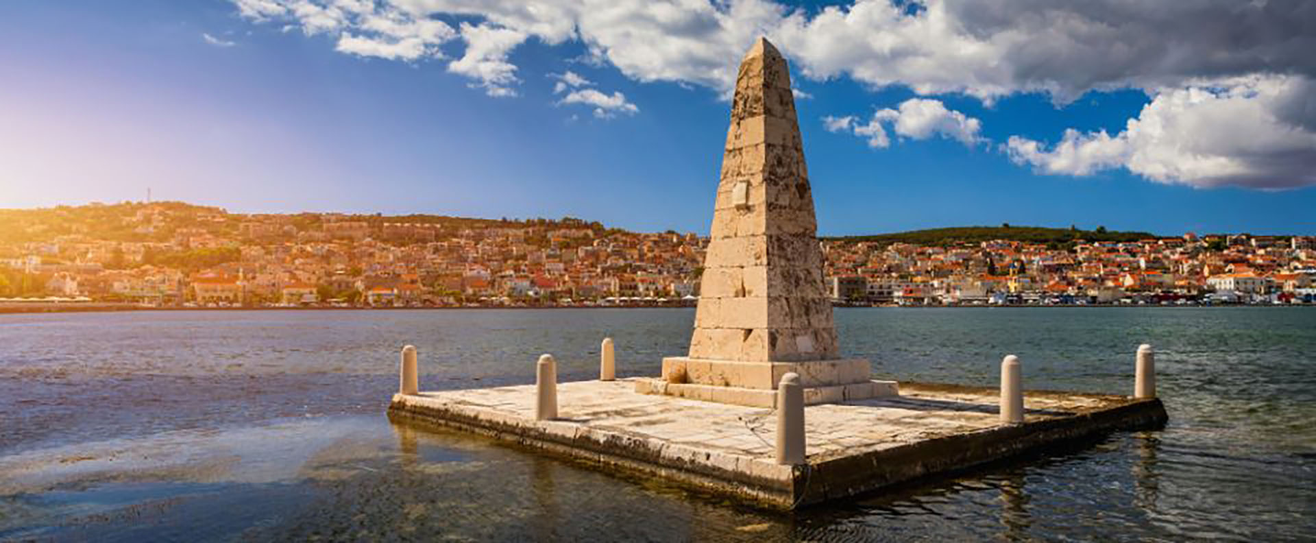 The Drapano Bridge Obelisk in Argostoli, Kefalonia, surrounded by calm waters and a vibrant coastal townscape, perfect for Ionian island adventures.