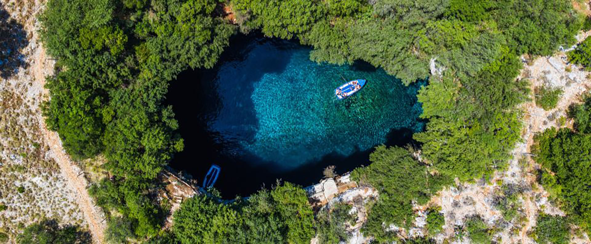 Aerial view of the Melissani Cave in Kefalonia, featuring crystal-clear turquoise waters surrounded by lush greenery, a hidden Ionian gem.