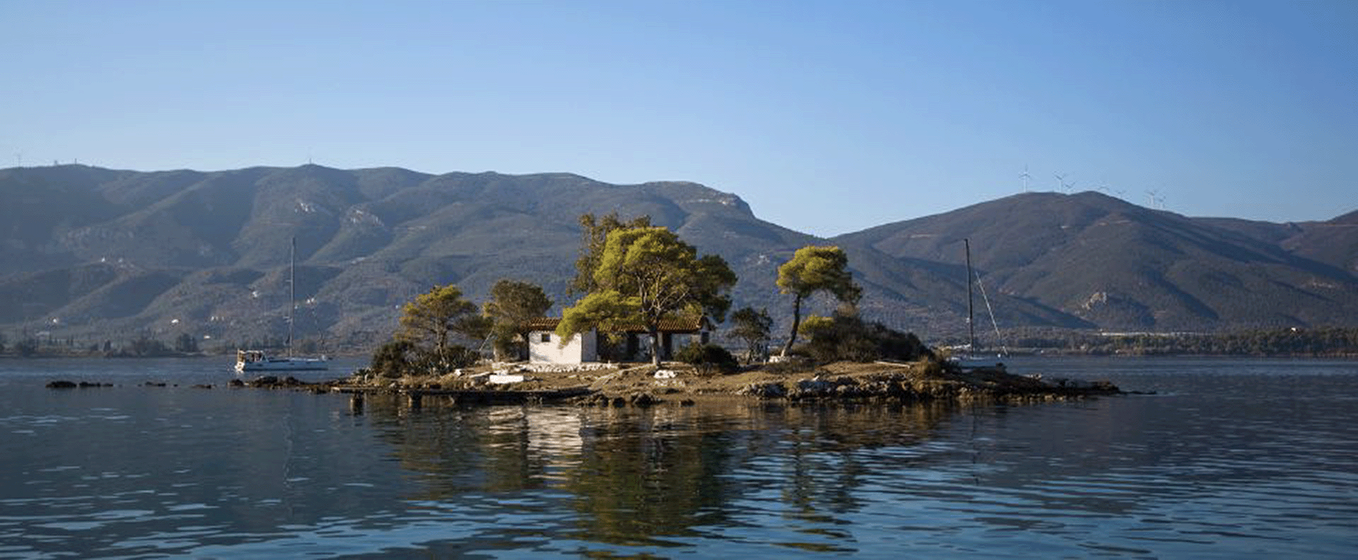 A tranquil islet in the Saronic Gulf surrounded by calm waters and framed by lush hills in the distance.