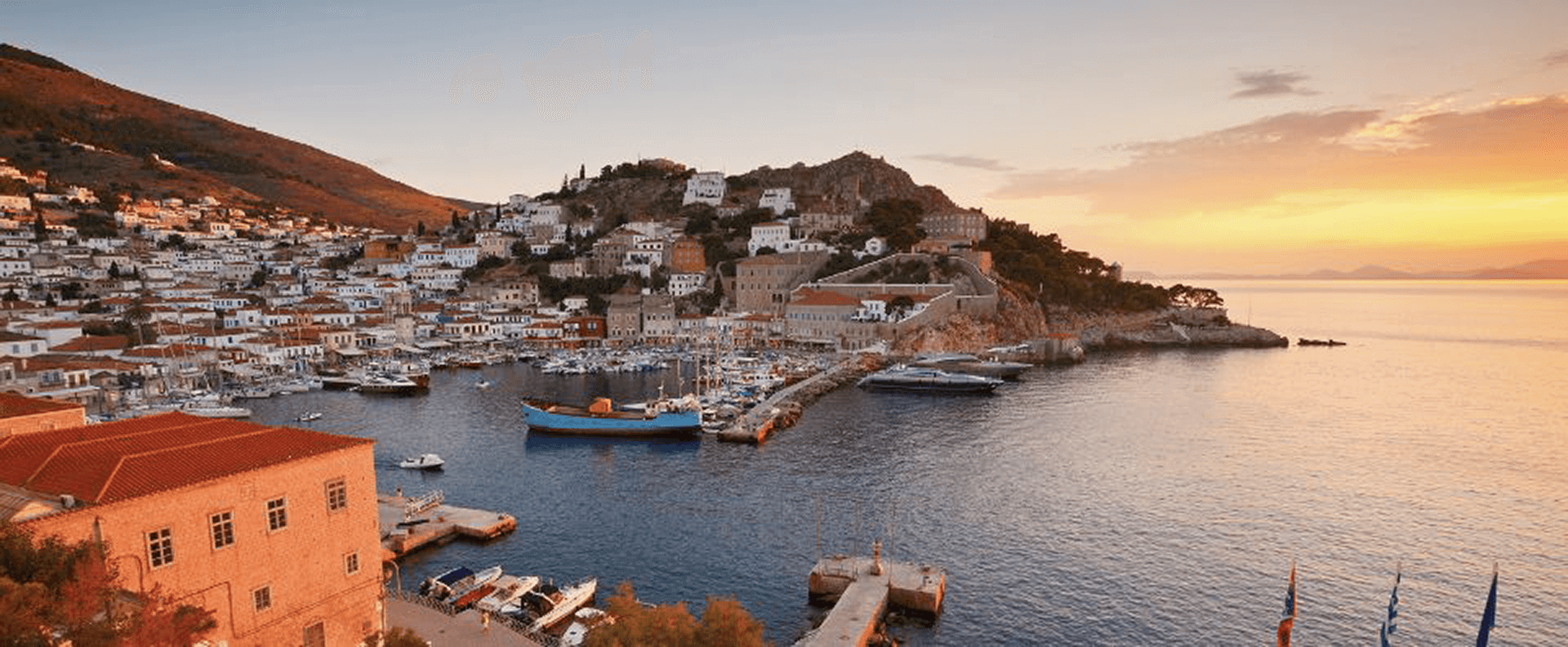 The picturesque harbour of Hydra in the Saronic Gulf Islands, featuring tranquil waters and a golden sunset over charming whitewashed houses.