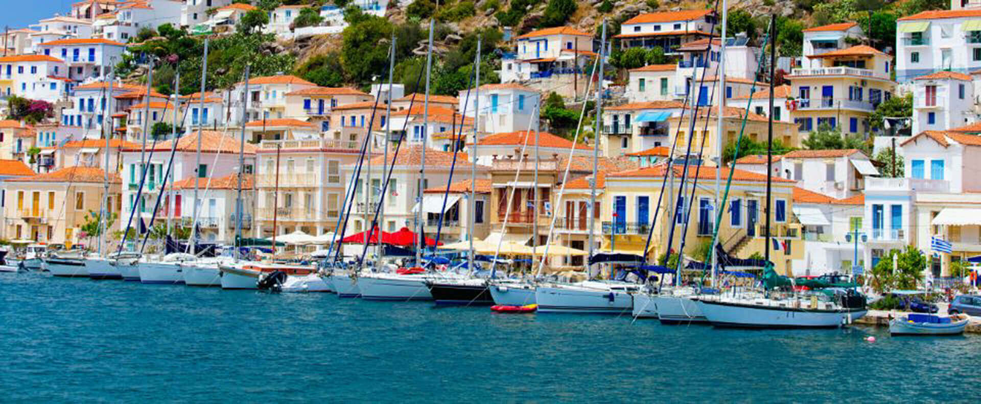 Sailing boats anchored along the vibrant waterfront of Poros, a serene destination in the Saronic Gulf Greece, with colourful houses in the background.