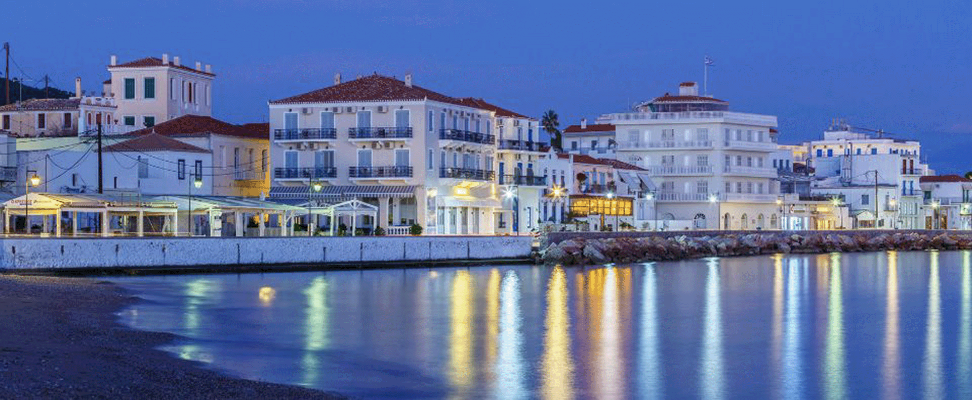 The evening glow reflects on the calm waters of Spetses harbour, showcasing elegant seaside mansions in the Saronic Gulf.