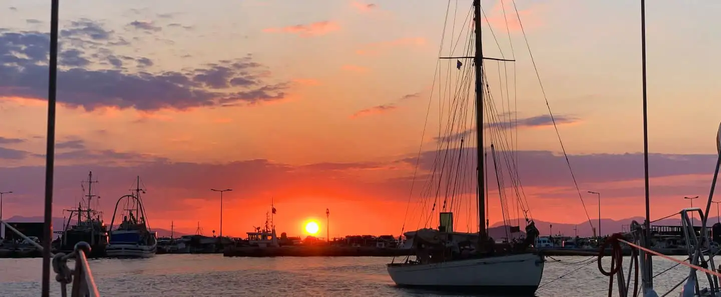 A sailboat moored in a Greek harbour at sunset, with warm orange and pink skies.