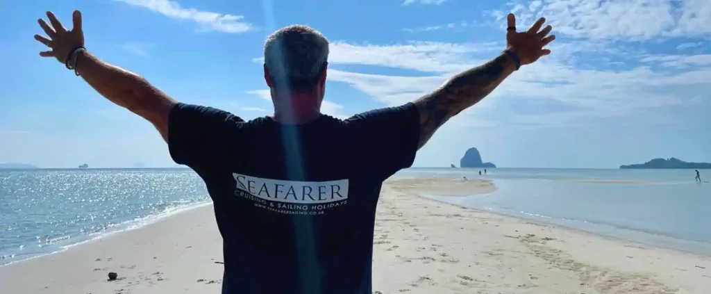 Man on a sandy beach with arms raised, facing the sea, wearing a Seafarer t-shirt—used in sailing blogs.