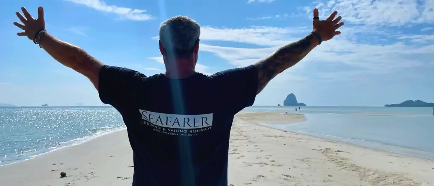 Man on a sandy beach with arms raised, facing the sea, wearing a Seafarer t-shirt—used in sailing blogs.