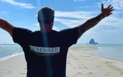 Man on a sandy beach with arms raised, facing the sea, wearing a Seafarer t-shirt—used in sailing blogs.