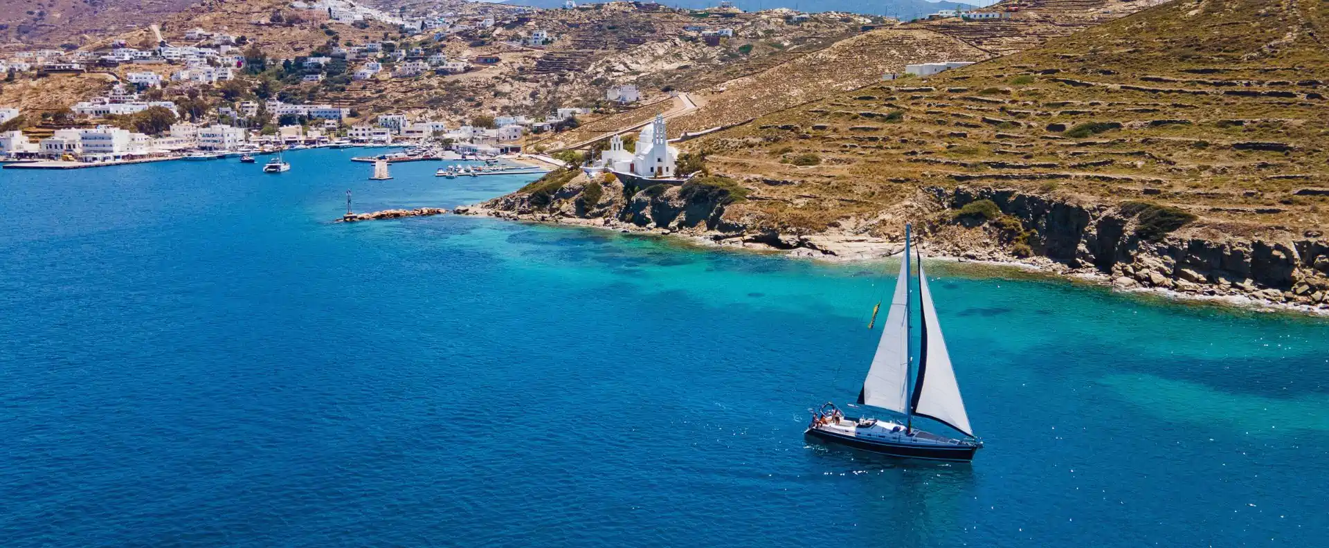 sailing blogs yacht near greek harbour coastline A sailing yacht featured in Seafarer’s sailing blogs gliding past a Greek island harbour and coastline.