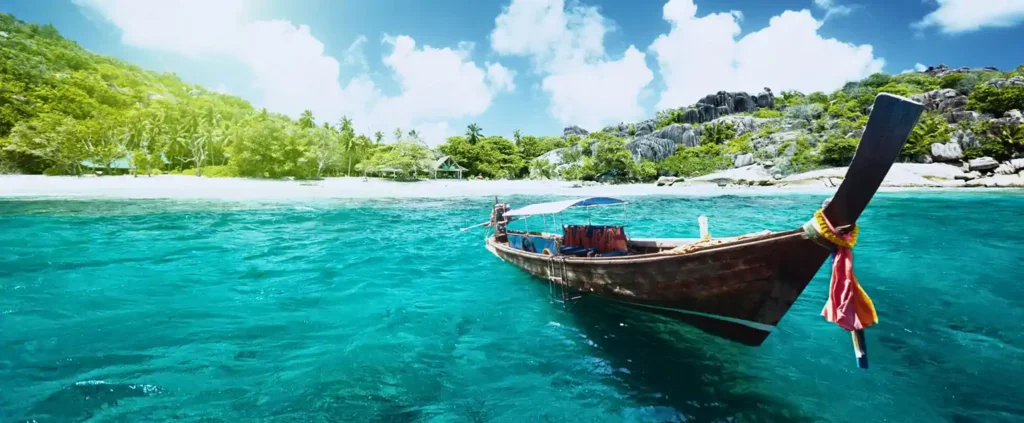 Traditional longtail boat floating near a tropical beach in Thailand with turquoise water, palm trees, and rocky coastline in the background