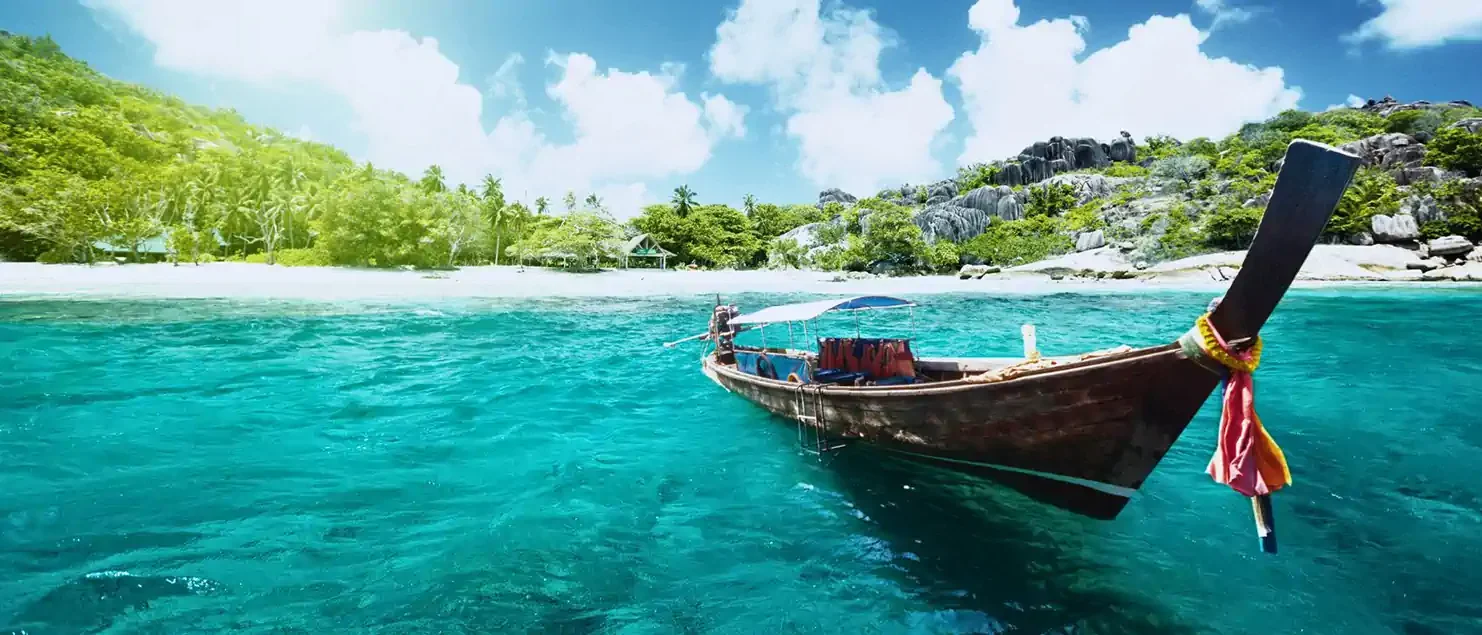 Traditional longtail boat floating near a tropical beach in Thailand with turquoise water, palm trees, and rocky coastline in the background