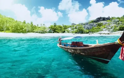 Traditional longtail boat floating near a tropical beach in Thailand with turquoise water, palm trees, and rocky coastline in the background