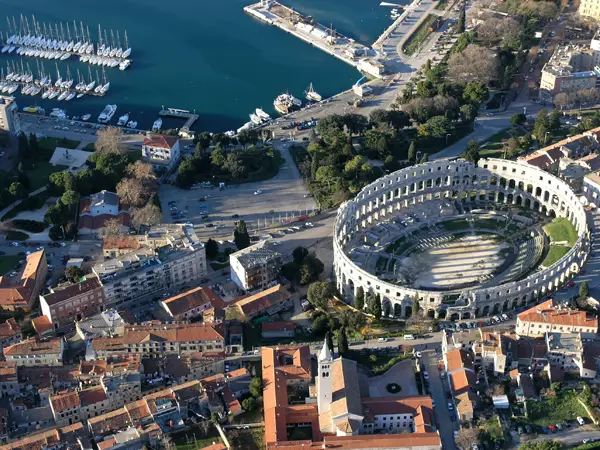 Aerial view of the historic coastal city of Pula featuring a large, well-preserved Roman amphitheater near the center, surrounded by dense red-roofed buildings, streets, and greenery, with a marina full of boats and deep blue water along the shoreline in the background.