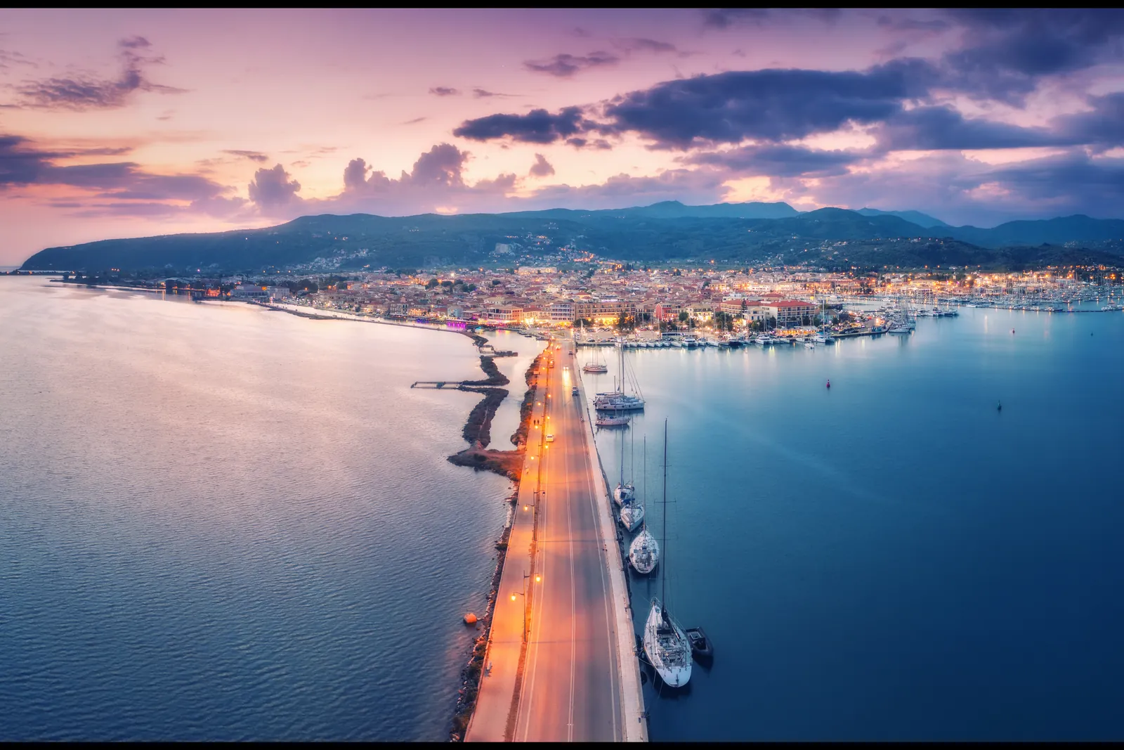 Aerial view of the Lefkas canal and causeway at dusk, with yachts moored below the lights of Lefkas town